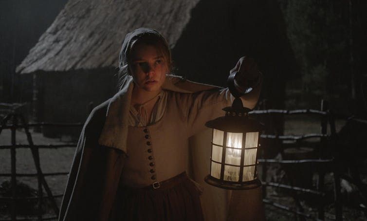 A woman dressed as a period holds a lamp in the middle of a village.